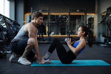 Sporty young couple exercising together in gym, man assisting fit lady while doing sits ups with...