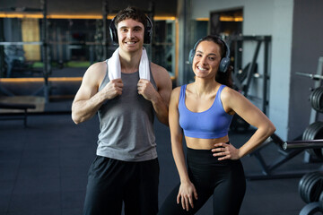 Fototapeta na wymiar Happy young woman and man in sportswear guy with towel on shoulders looking and smiling at camera together, posing in modern gym