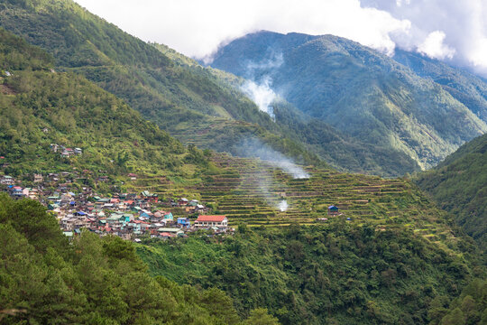 A scenic view of a remote mountain village amid terraced fields, with clouds drifting over green hills. At the Bay-yo Rice Terraces, near the town of Bontoc, Mountain Province.