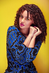 Medium shot of a woman with curly hair, blue dress, makeup, and yellow background