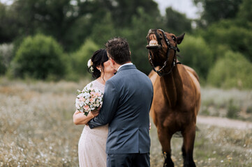Funny horse laughs showing teeth while standing with bride and groom hugging and kissing outdoors....