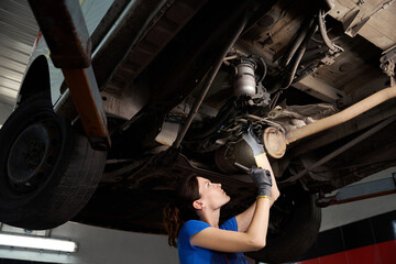 Female inspects chassis of car, raised on a lift - obrazy, fototapety, plakaty