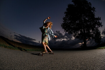 Fototapeta premium Young smiling girl in a dress and a denim jacket is riding a longboard on an asphalt road