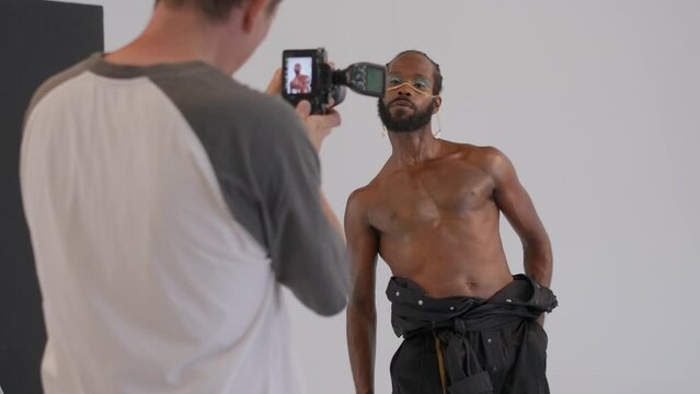 Professional photographer taking pictures of shirtless muscular African American gay man with makeup in studio on white background