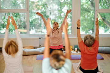 A group of senior women engage in various yoga exercises, including neck, back, and leg stretches, under the guidance of a trainer in a sunlit space, promoting well-being and harmony