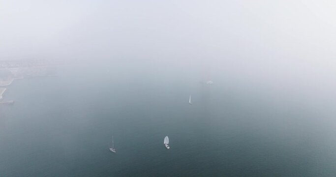 Aerial drone shot of sailing yacht boats sailing in thick mist and fog by the 25th April Bridge in Lisbon, Portugal, Europe. Shot in ProRes 422 HQ