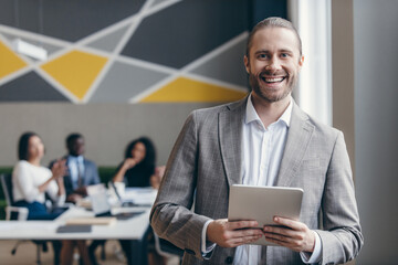 Smiling young businessman holding digital tablet while his colleagues working on background