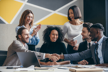 Two confident businessmen shaking hands while sitting with their colleagues at the office desk