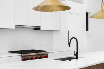 A kitchen detail with a gold light fixtures hanging over a white marble island and black faucet,...