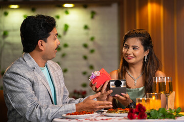 Joyful happy woman giving watch as gift to husband during anniversary or birthday celebration at candlelight dinner - concept of loving couples, surprise present and special occasion.