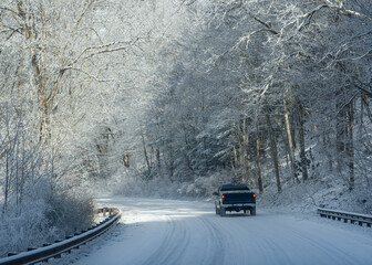 A lone pickup truck drives through a wintry mountain landscape on a snow-covered road. The road curves ahead with guardrails on both sides. 