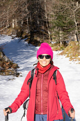 photograph of woman in the forest with walking sticks and backpack. Snow and trees. look at camera