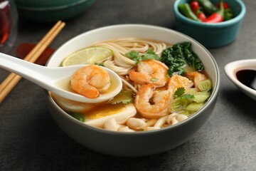 Eating delicious ramen with shrimps from bowl with spoon at grey table, closeup. Noodle soup