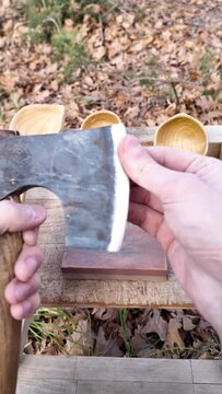 Close-up of a carpenter checks the sharpness of an axe and starts to sharpen it. He sits on a camping chair in a forest.