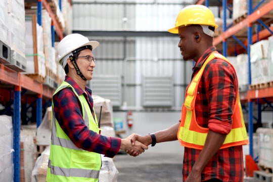 Happy Asian senior supervisor wearing safety vest and helmet, shaking hands with his colleague worker at cargo logistics warehouse. African engineer man having hand shack with elderly Asian worker.