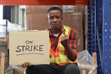 Angry unhappy African worker man wearing safety vest and giving the middle finger with strike banner placard sign at cargo logistic warehouse. Striking worker protesting at workplace. - obrazy, fototapety, plakaty