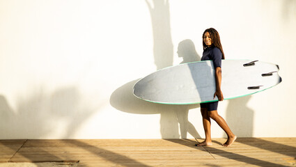A woman walks near a wall with a surfboard dressed in a wetsuit. Concept of selling surfboards, advertising surfing equipment. Copy space for water sports ads.