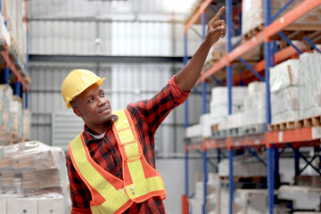Young African worker man wearing safety vest and helmet, standing and pointing at warehouse...