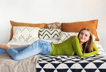 Young smiling positive teen girl in casual clothes lying on bed in her cozy room at home