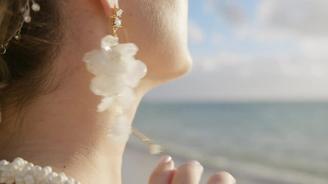 Close up side view of beautiful woman wearing luxury hanging white flower earrings. Side view of bride slowly touching boho style floral drop earrings. Girls hand with wedding diamond ring at beach 4K