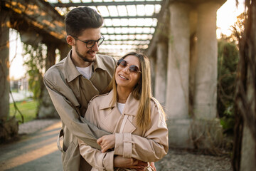 Portrait of a girl and a guy posing against the backdrop of an arch in the garden at sunset in spring. Family photo of a stylish couple in beige trench coats.
