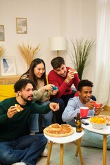 Group of multiethnic friends eating pizza and playing video games in shared apartment.