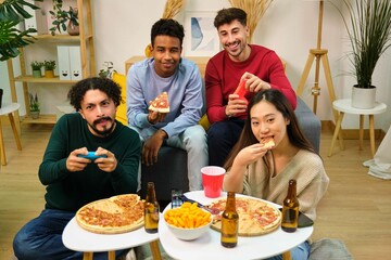 Group of multiethnic friends eating pizza and playing video games in shared apartment.
