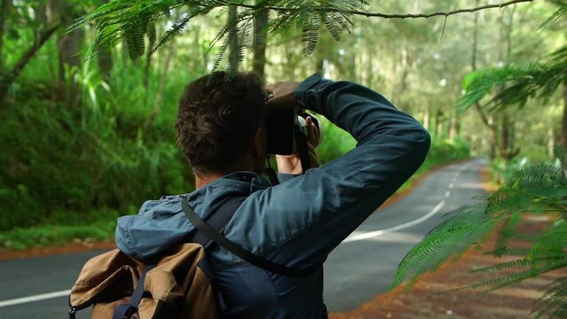Active adventure middle age man travel photographer with backpack shooting by camera beautiful forest landscape with asphalt road. Back view.