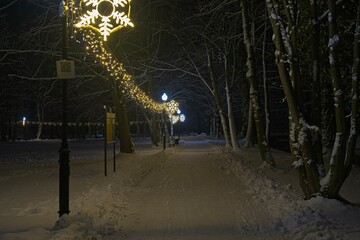 Zimowy wieczór w parku. Parkowa alejka pokryta warstwą białego śniegu. Z prawej strony znajduje się girlanda  świetlna rozświetlająca ciemności.