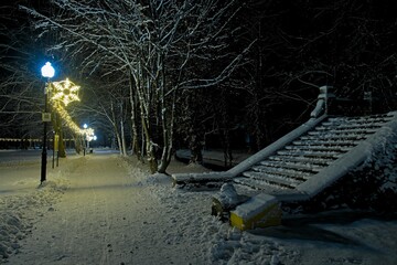 Zimowy wieczór w parku. Parkowa alejka pokryta warstwą białego śniegu. Z prawej strony znajduje się girlanda  świetlna rozświetlająca ciemności.