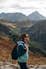 Stylish young woman with backpack and sunglasses standing on top of a mountain and looking to the Tatra rocks, Slovakia