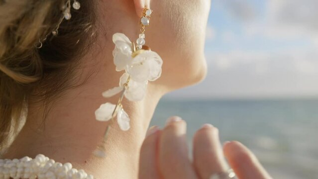 The girl hand straightens drop flower earrings. Young woman checking elegant hanging floral earring before boho wedding ceremony at beach. Bride wearing boho style accessories. Diamond ring jewelry 4K