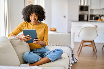 Happy young African American woman sitting on couch at home using digital tablet. Smiling relaxed...