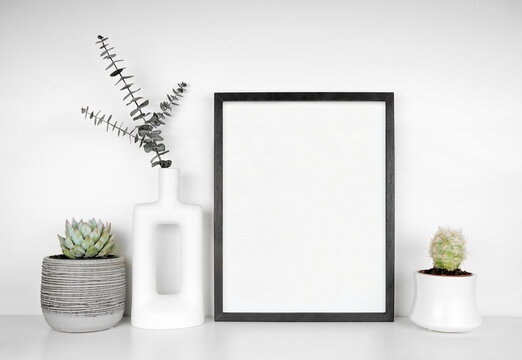 Mock up black frame with plants and eucalyptus in a modern vase. White shelf against a white wall. Copy space.