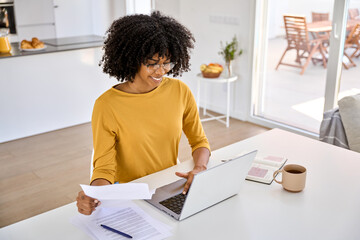 Young smiling happy African American woman checking financial paper calculating banking loan or household payments using laptop computer paying bills online sitting at home table in kitchen. - obrazy, fototapety, plakaty