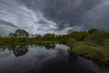 The river reflects the dramatic sky, a spring motif with the river and green bushes and grass.