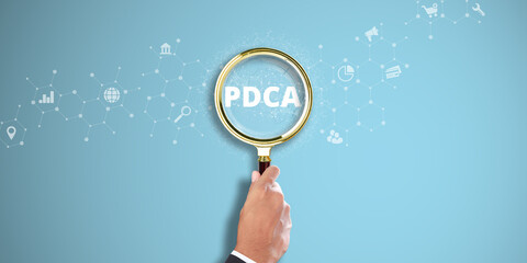 PDCA: Businessman Hand Holding a Magnifying Glass with Plan-Do-Check-Act (PDCA) Icon on Light Blue Background. Continuous Improvement, Quality Assurance, Process Optimization.