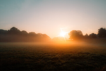 a beatuful countryside sunrise / sunset over agricultural farm land. sun flare producing mist on a dewy grassy field lined with trees. No people with copy space, web design wallpaper / background