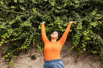 Carefree woman with arms raised standing against ivy wall in park