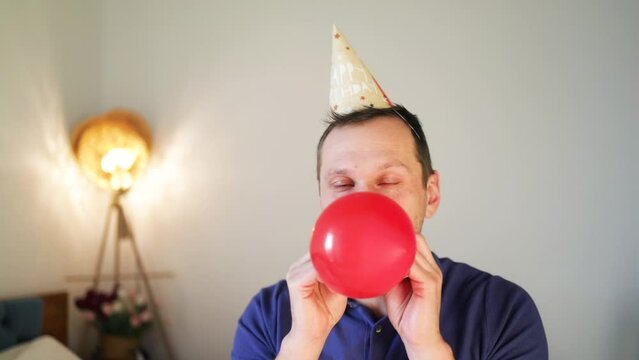 Young Caucasian man in birthday cap inflating red ballon sitting on sofa. The concept of the holiday. 