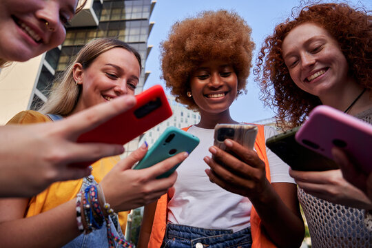 Low angle view of a group of multiracial young women friends using smartphones addicted to technology. People gathered in a circle immersed in mobiles. Social media communication.