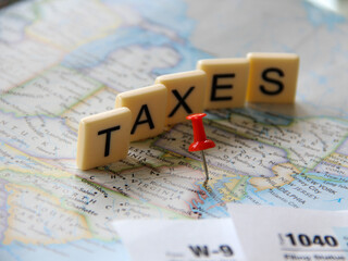 Selective focus of red push pin tack pining Washington DC in a map with the word Taxes in letter tiles in background and tax forms in foreground for tax returns filing concept due in April.