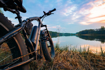 Electric bike with thick wheels on the background of lake and sunset. Sports fatbike. Picturesque place in the village. The concept of a healthy lifestyle.