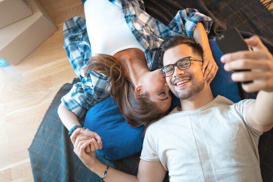 Happy young couple moving in new house and taking self portrait with smart phone. Young man and woman lying on floor making selfie with mobile phone.