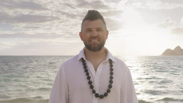 Handsome male person in boho wedding outfit with traditional kukui nut necklace, Hawaii islands. Charming man looking at camera at beach. Portrait smiling groom in white linen shirt and stylish hairdo