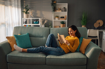 Obraz premium Young woman lying on comfortable sofa reading a book in her free time at home