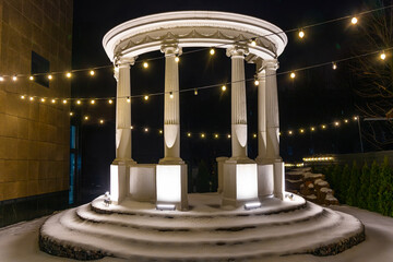A beautiful white gazebo with columns in an antique style, illuminated by electric lamps. Vintage...