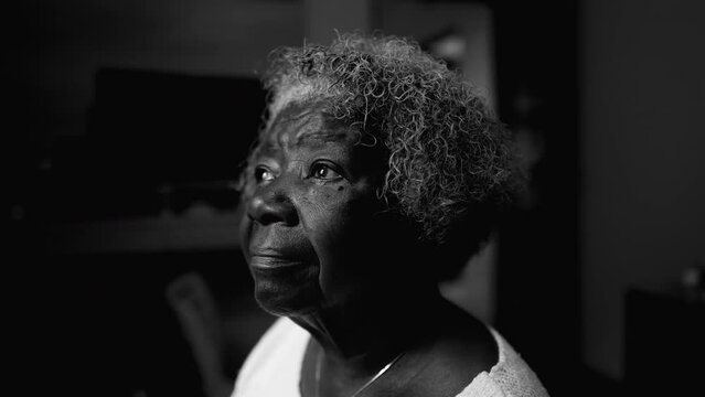 One introspective black senior woman in solitude at home in black and white. Monochromatic close-up face of an African American lady staring at window with thoughtful emotion