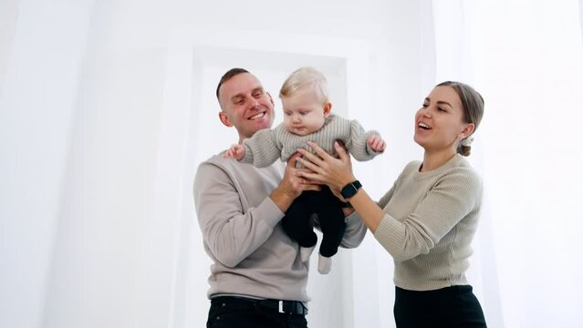 Smiling Caucasian couple holding a little baby from two sides. Parents wave their infant son. White backdrop.