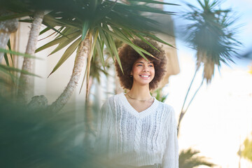 a young latina woman with afro hair smiling happily among plants in a plaza